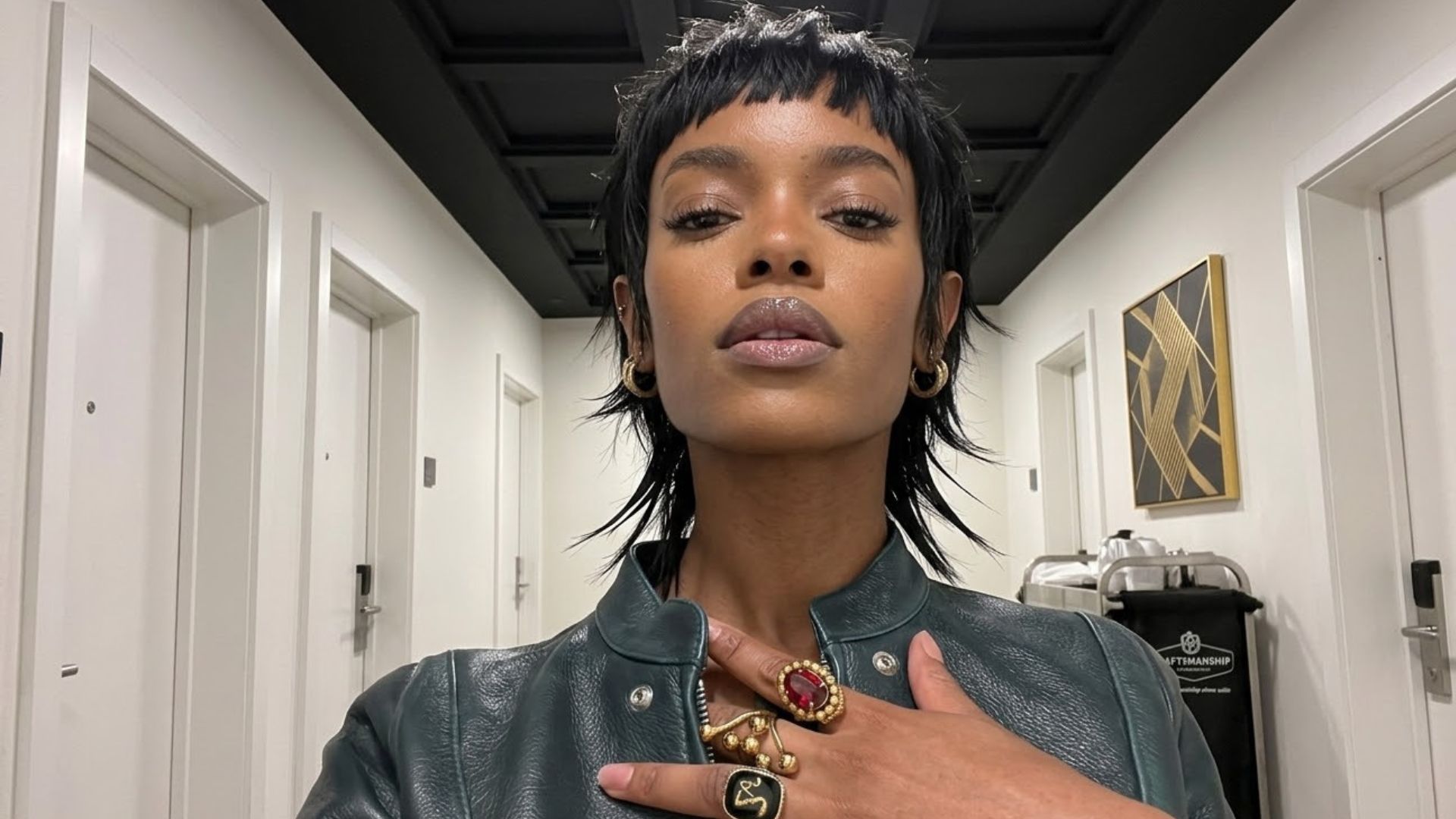 Un selfie iperrealistico a bassa angolazione di una donna afroamericana con un taglio di capelli 'shaggy wolf-cut' (mullet strutturato) nero e testurizzato, che indossa una giacca di pelle verde scuro. Ha la mano sul petto, mostrando un grande anello d'oro oversize con una gemma rossa ovale e altri anelli con simboli neri. Si trova in un corridoio d'hotel minimalista con porte bianche identiche.