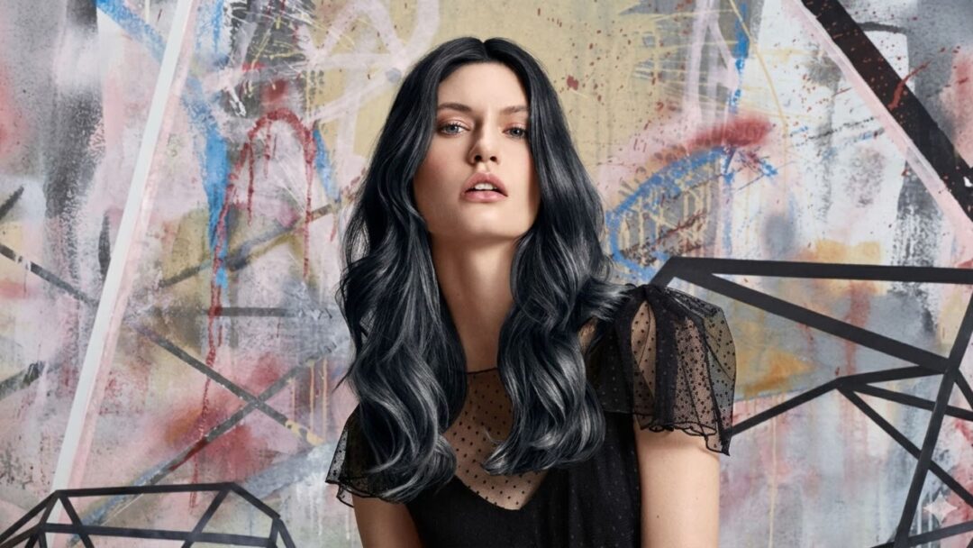 Una modella con lunghi capelli ondulati grigio-argento e neri, in un abito nero con spalle a pois traslucide, posa davanti a un murale urbano geometrico e colorato.