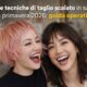 Due modelle sorridenti in salone mostrano un taglio scalato moderno per la primavera 2026, capelli texturizzati e movimento naturale.