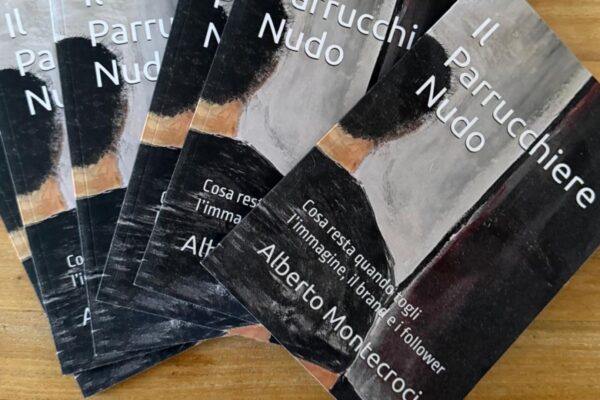 Una foto di un ventaglio di circa sette copie del libro cartaceo 'Il Parrucchiere Nudo' dell'autore Alberto Montecroci, disposte su un tavolo di legno chiaro con venature visibili. La copia a destra è più a fuoco e mostra il titolo completo e l'autore. La copertina ha una grafica scura ed espressionista di un uomo visto da dietro.