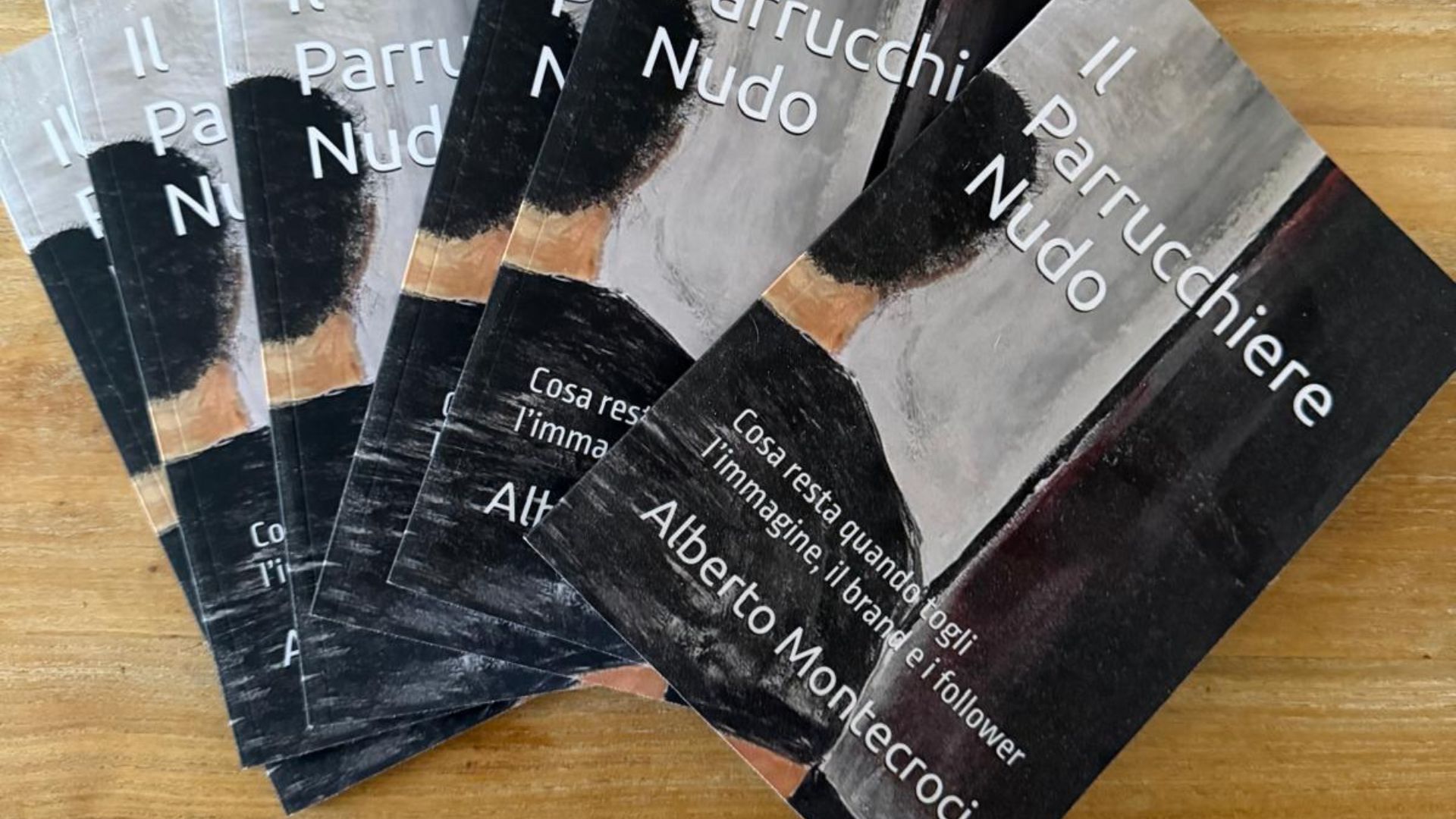 Una foto di un ventaglio di circa sette copie del libro cartaceo 'Il Parrucchiere Nudo' dell'autore Alberto Montecroci, disposte su un tavolo di legno chiaro con venature visibili. La copia a destra è più a fuoco e mostra il titolo completo e l'autore. La copertina ha una grafica scura ed espressionista di un uomo visto da dietro.