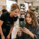 A blonde hair stylist wearing a black apron with the "ARMONIA" logo is leaning over to look at a green iPhone held by a brunette client who is sitting in a salon chair and wearing a dark grey drape. They are both looking at the screen with interested expressions. The background is a busy professional hair salon with other stylists, clients, and product shelves.