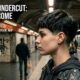 Modella con taglio corto Nape Undercut tecnico, vista laterale e posteriore in una stazione metropolitana underground di Roma. La nuca presenta una sfumatura millimetrica di precisione con tatuaggi minimalisti. Atmosfera urban ed editoriale.