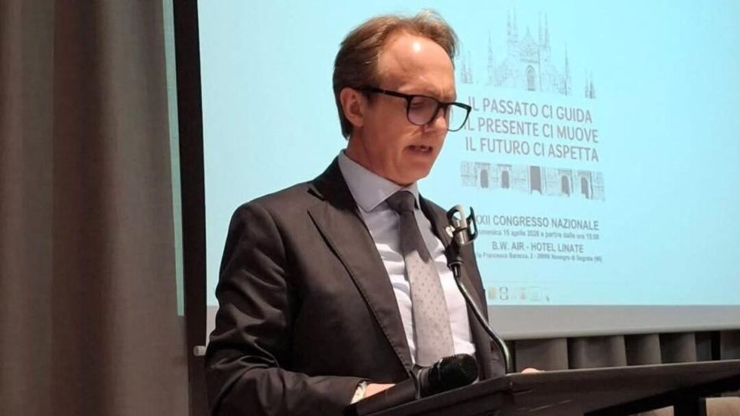 Un uomo con occhiali e completo parla al podio durante un congresso nazionale a Milano. Dietro di lui, uno schermo proietta una slide con l'immagine del Duomo di Milano e il testo "IL PASSATO CI GUIDA. IL PRESENTE CI MUOVE. IL FUTURO CI ASPETTA. XII CONGRESSO NAZIONALE".