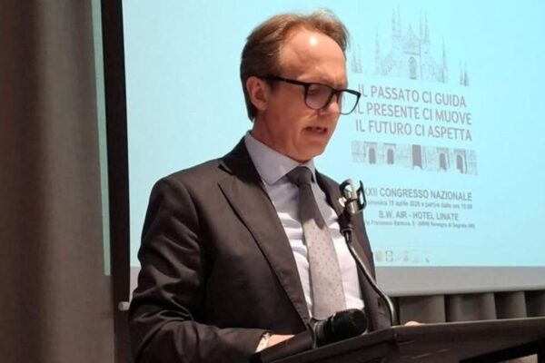 Un uomo con occhiali e completo parla al podio durante un congresso nazionale a Milano. Dietro di lui, uno schermo proietta una slide con l'immagine del Duomo di Milano e il testo "IL PASSATO CI GUIDA. IL PRESENTE CI MUOVE. IL FUTURO CI ASPETTA. XII CONGRESSO NAZIONALE".
