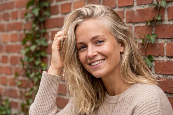 Primo piano di una modella con tecnica Scandinavian Hairline su capelli biondi, posa naturale davanti a un muro di mattoni rossi.