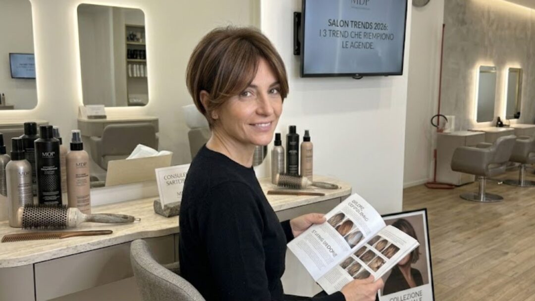 Donna over 40 sorridente con taglio capelli bixie castano e schiariture face framing, seduta in un salone di parrucchiere moderno mentre consulta una rivista di moda capelli MDP.