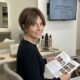 Donna over 40 sorridente con taglio capelli bixie castano e schiariture face framing, seduta in un salone di parrucchiere moderno mentre consulta una rivista di moda capelli MDP.