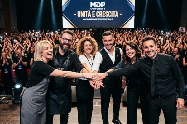 Un gruppo di sei parrucchieri e leader del settore hair & beauty unisce le mani sul palco principale del MDP Congress in un gesto di unità. Sullo sfondo, un grande schermo a LED mostra il testo "- UNITÀ E CRESCITA -" e un auditorium immenso e gremito di migliaia di colleghi applaude e scatta foto.