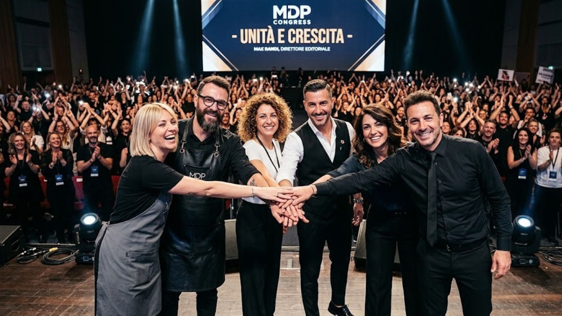 Un gruppo di sei parrucchieri e leader del settore hair & beauty unisce le mani sul palco principale del MDP Congress in un gesto di unità. Sullo sfondo, un grande schermo a LED mostra il testo "- UNITÀ E CRESCITA -" e un auditorium immenso e gremito di migliaia di colleghi applaude e scatta foto.