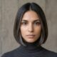 Front-facing half-length photographic portrait of a contemporary woman with a dark bob haircut featuring distinct grey and silver highlights, wearing a dark grey turtleneck, against a textured concrete wall.