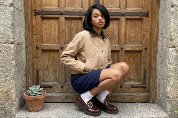 Una giovane donna con un taglio a caschetto nero è accovacciata su un gradino di pietra. Indossa una giacca beige stile bomber, una gonna blu scuro testurizzata, calzini bianchi e mocassini bordeaux chunky con fibbie dorate. Dietro di lei c'è un'imponente porta di legno antica e borchiata. A sinistra si vede una pianta succulenta in vaso.