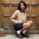 Una giovane donna con un taglio a caschetto nero è accovacciata su un gradino di pietra. Indossa una giacca beige stile bomber, una gonna blu scuro testurizzata, calzini bianchi e mocassini bordeaux chunky con fibbie dorate. Dietro di lei c'è un'imponente porta di legno antica e borchiata. A sinistra si vede una pianta succulenta in vaso.