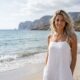 Una donna bionda sorridente in un abito di lino bianco su una spiaggia sabbiosa in riva al mare con scogliere sullo sfondo.
