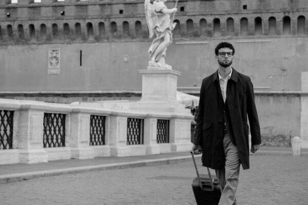 Uomo con occhiali, camicia e soprabito scuro cammina sul ponte di Castel Sant'Angelo a Roma trascinando un trolley nero, in un'immagine in bianco e nero con una statua d'angelo sullo sfondo.