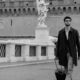 Uomo con occhiali, camicia e soprabito scuro cammina sul ponte di Castel Sant'Angelo a Roma trascinando un trolley nero, in un'immagine in bianco e nero con una statua d'angelo sullo sfondo.