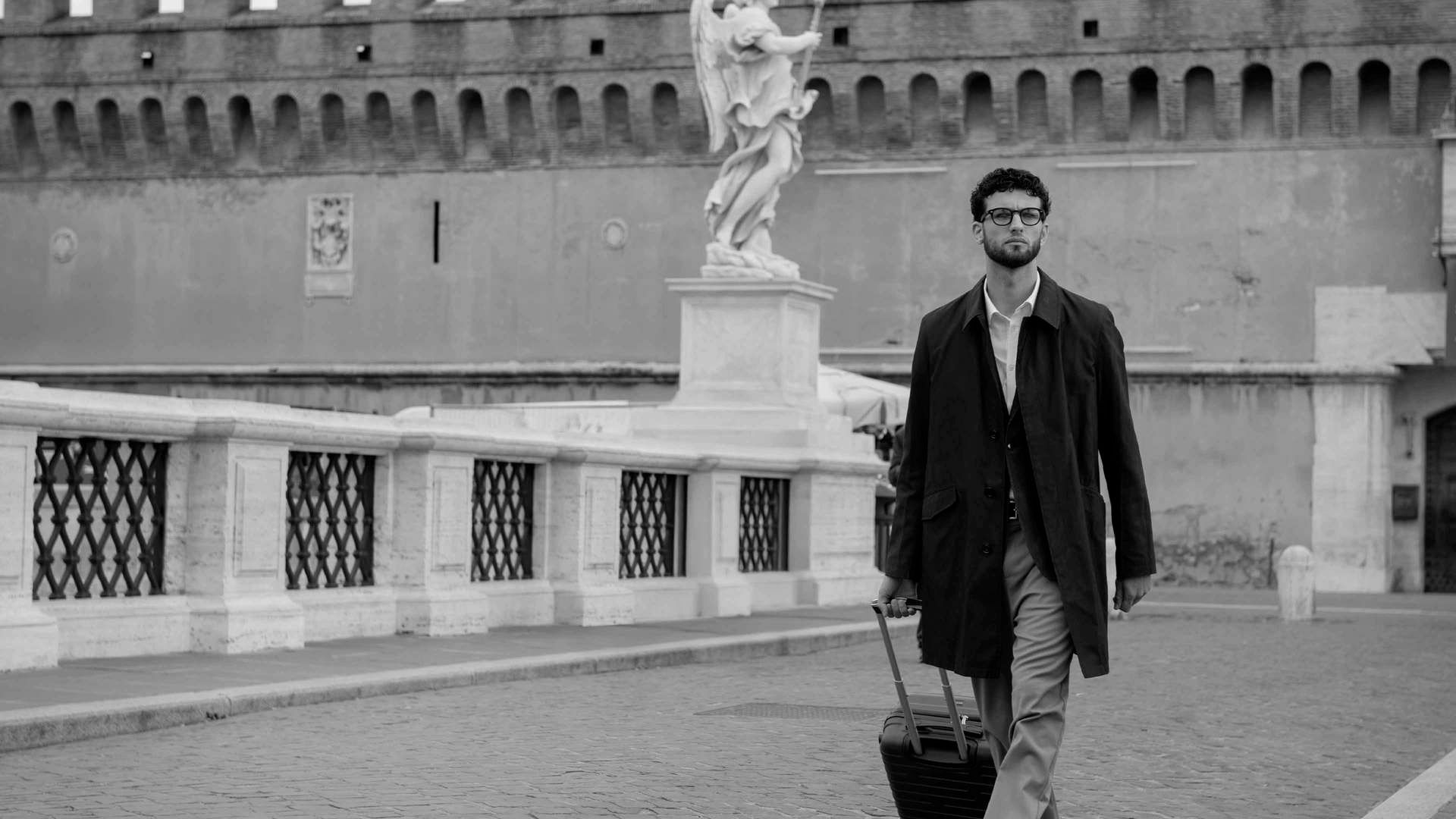 Uomo con occhiali, camicia e soprabito scuro cammina sul ponte di Castel Sant'Angelo a Roma trascinando un trolley nero, in un'immagine in bianco e nero con una statua d'angelo sullo sfondo.