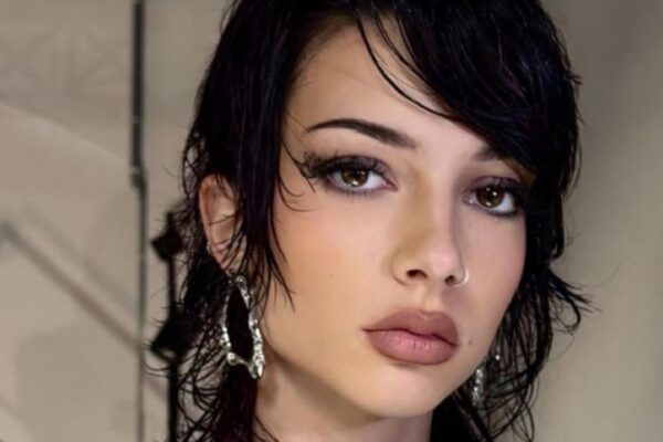 Primo piano di una modella con un taglio corto e disordinato di colore scuro, stile mullet shag bagnato, trucco intenso e piercing al naso. Indossa un abito nero strutturato, avant-garde. Sfondo backstage industriale con tralicci e pareti in cemento.
