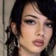 Primo piano di una modella con un taglio corto e disordinato di colore scuro, stile mullet shag bagnato, trucco intenso e piercing al naso. Indossa un abito nero strutturato, avant-garde. Sfondo backstage industriale con tralicci e pareti in cemento.