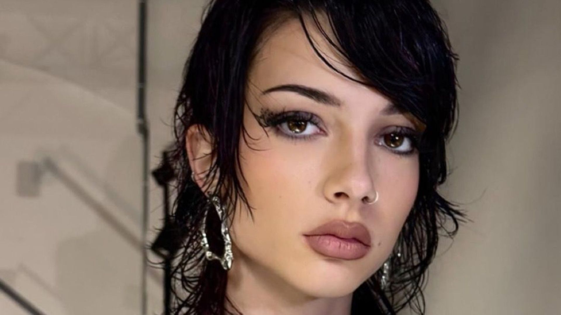 Primo piano di una modella con un taglio corto e disordinato di colore scuro, stile mullet shag bagnato, trucco intenso e piercing al naso. Indossa un abito nero strutturato, avant-garde. Sfondo backstage industriale con tralicci e pareti in cemento.