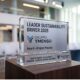 Trofeo in vetro e metallo del premio Leader Sustainability Driver 2026 assegnato al Gruppo Ymenso, posizionato su un piedistallo in legno con una sala conferenze sfocata sullo sfondo.
