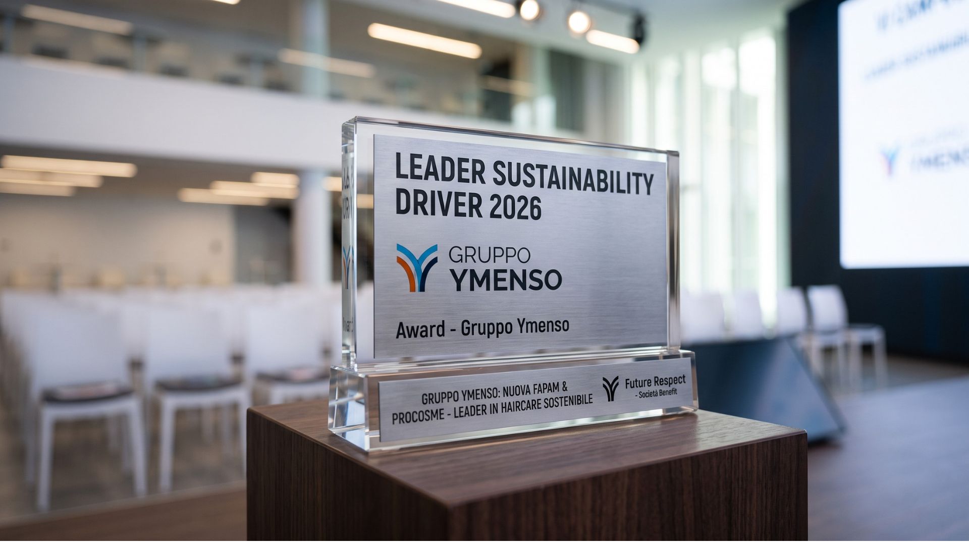 Trofeo in vetro e metallo del premio Leader Sustainability Driver 2026 assegnato al Gruppo Ymenso, posizionato su un piedistallo in legno con una sala conferenze sfocata sullo sfondo.