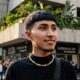 Ragazzo giovane con taglio di capelli stile Edgar cut, frangia geometrica dritta e sfumatura laterale alta, indossa una collana di perle colorate e un lupetto nero.