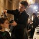 Hairstylist in completo nero acconcia i capelli di una modella durante un backstage in una sala storica con lampadario di cristallo