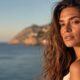 Donna con capelli mossi castani illuminati dalla luce dorata del tramonto, sullo sfondo il mare e una costa rocciosa mediterranea