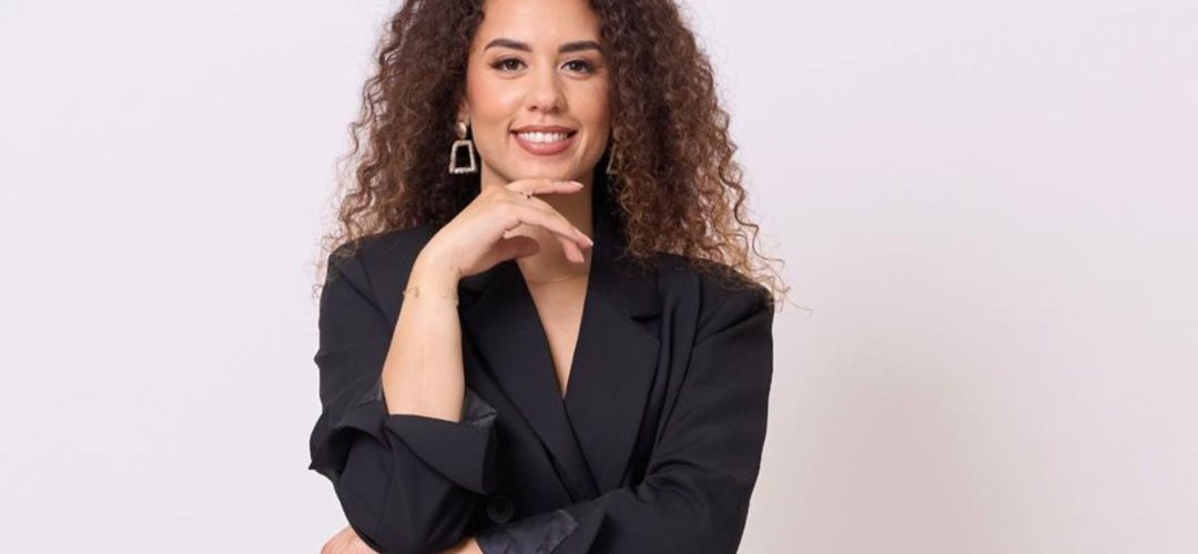 Ritratto professionale di una giovane donna con capelli ricci e castani, che sorride con sicurezza indossando un blazer nero su sfondo bianco.