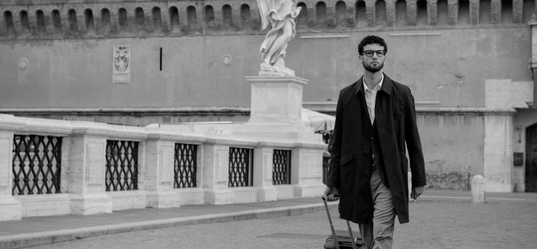 Uomo con occhiali, camicia e soprabito scuro cammina sul ponte di Castel Sant'Angelo a Roma trascinando un trolley nero, in un'immagine in bianco e nero con una statua d'angelo sullo sfondo.