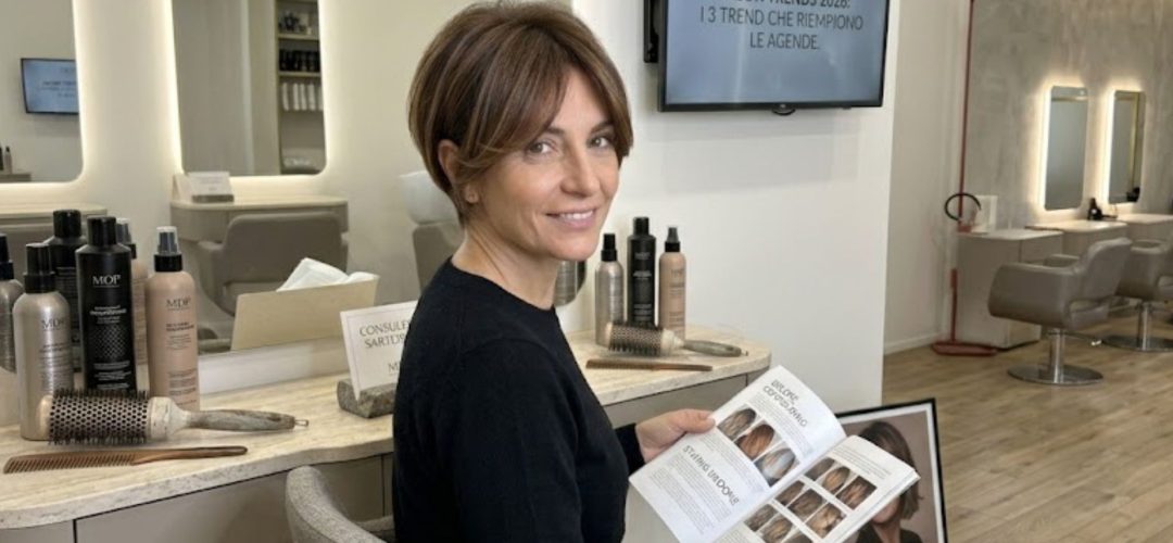 Donna over 40 sorridente con taglio capelli bixie castano e schiariture face framing, seduta in un salone di parrucchiere moderno mentre consulta una rivista di moda capelli MDP.
