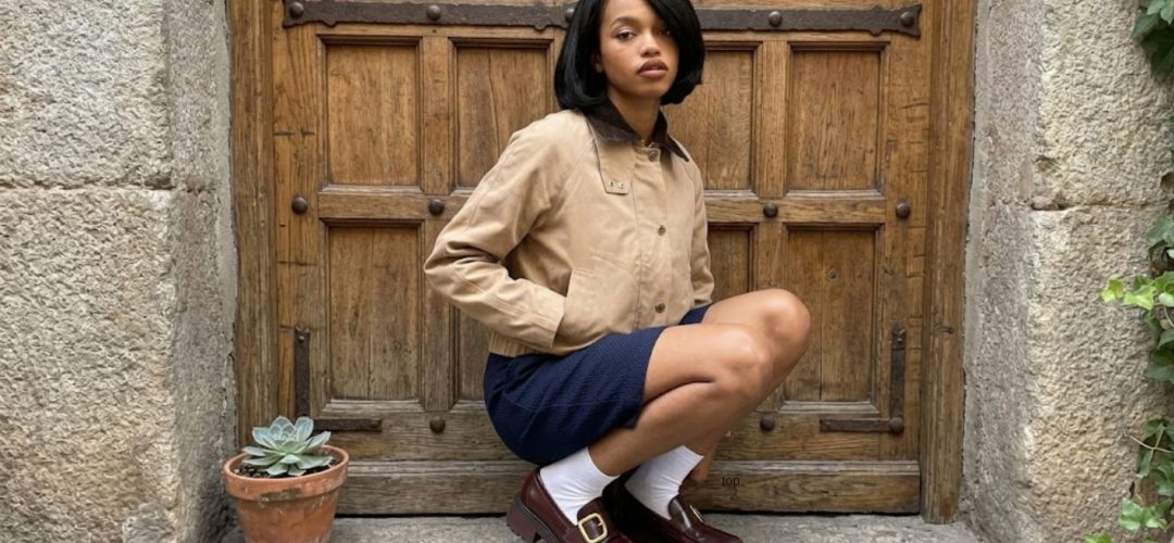 Una giovane donna con un taglio a caschetto nero è accovacciata su un gradino di pietra. Indossa una giacca beige stile bomber, una gonna blu scuro testurizzata, calzini bianchi e mocassini bordeaux chunky con fibbie dorate. Dietro di lei c'è un'imponente porta di legno antica e borchiata. A sinistra si vede una pianta succulenta in vaso.