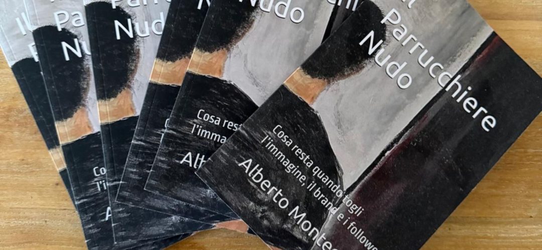 Una foto di un ventaglio di circa sette copie del libro cartaceo 'Il Parrucchiere Nudo' dell'autore Alberto Montecroci, disposte su un tavolo di legno chiaro con venature visibili. La copia a destra è più a fuoco e mostra il titolo completo e l'autore. La copertina ha una grafica scura ed espressionista di un uomo visto da dietro.