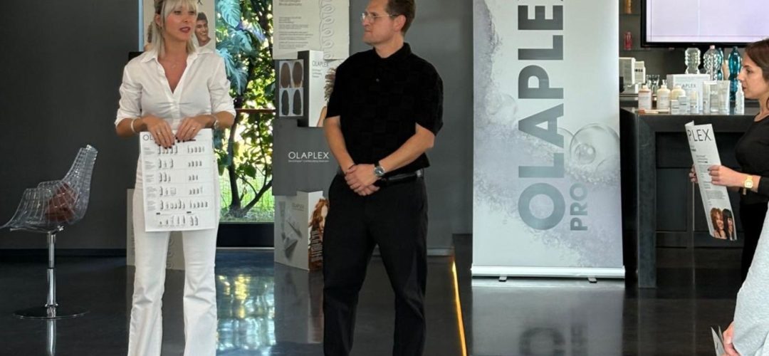 La formatrice Francesca Ventura in abito bianco presenta un foglio tecnico durante un seminario Olaplex in collaborazione con Academy Calenda, affiancata da un collega in abito nero.