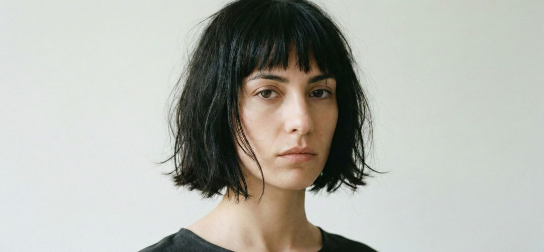 Ritratto frontale di giovane donna con taglio di capelli a caschetto nero stile Japanese Bob spettinato, frangia irregolare e look naturale su sfondo neutro.