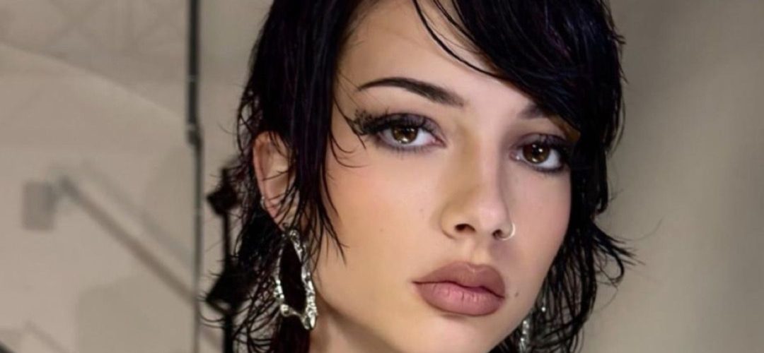 Primo piano di una modella con un taglio corto e disordinato di colore scuro, stile mullet shag bagnato, trucco intenso e piercing al naso. Indossa un abito nero strutturato, avant-garde. Sfondo backstage industriale con tralicci e pareti in cemento.