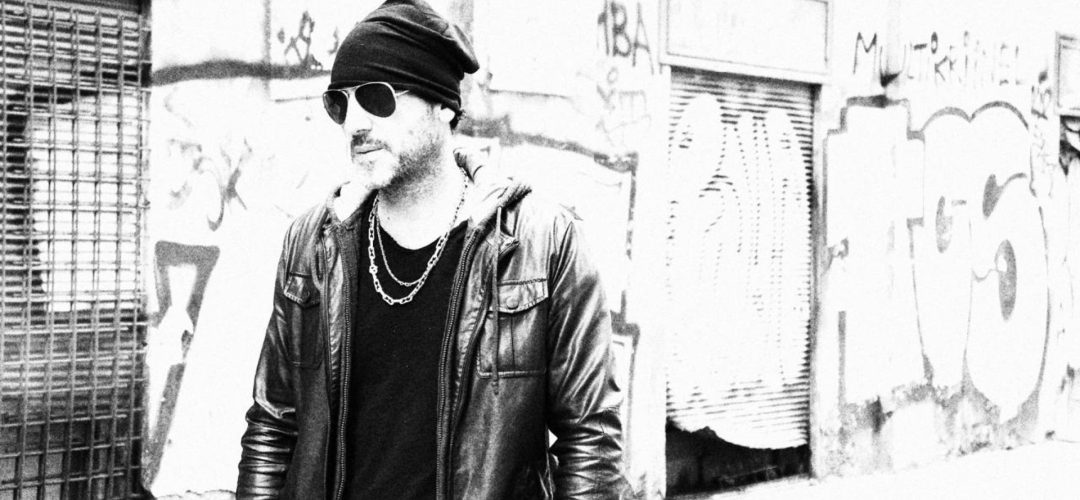 Ritratto in bianco e nero di Antonio Tufano, Hair Architect, in stile urban con giacca di pelle e occhiali da sole, posato di fronte a un muro con graffiti.