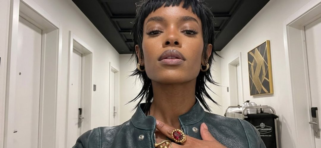 Un selfie iperrealistico a bassa angolazione di una donna afroamericana con un taglio di capelli 'shaggy wolf-cut' (mullet strutturato) nero e testurizzato, che indossa una giacca di pelle verde scuro. Ha la mano sul petto, mostrando un grande anello d'oro oversize con una gemma rossa ovale e altri anelli con simboli neri. Si trova in un corridoio d'hotel minimalista con porte bianche identiche.