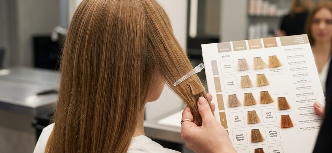 Primo piano delle mani di un parrucchiere che accosta una cartella colori a una ciocca di capelli biondo scuro di una cliente per scegliere la giusta nuance in salone.