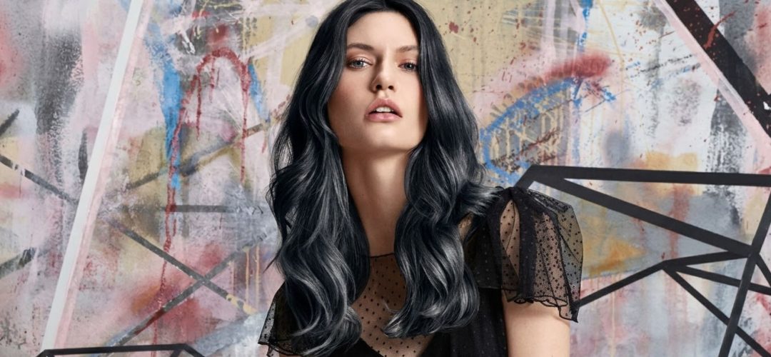 Una modella con lunghi capelli ondulati grigio-argento e neri, in un abito nero con spalle a pois traslucide, posa davanti a un murale urbano geometrico e colorato.