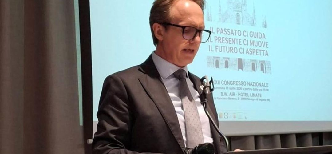 Un uomo con occhiali e completo parla al podio durante un congresso nazionale a Milano. Dietro di lui, uno schermo proietta una slide con l'immagine del Duomo di Milano e il testo "IL PASSATO CI GUIDA. IL PRESENTE CI MUOVE. IL FUTURO CI ASPETTA. XII CONGRESSO NAZIONALE".