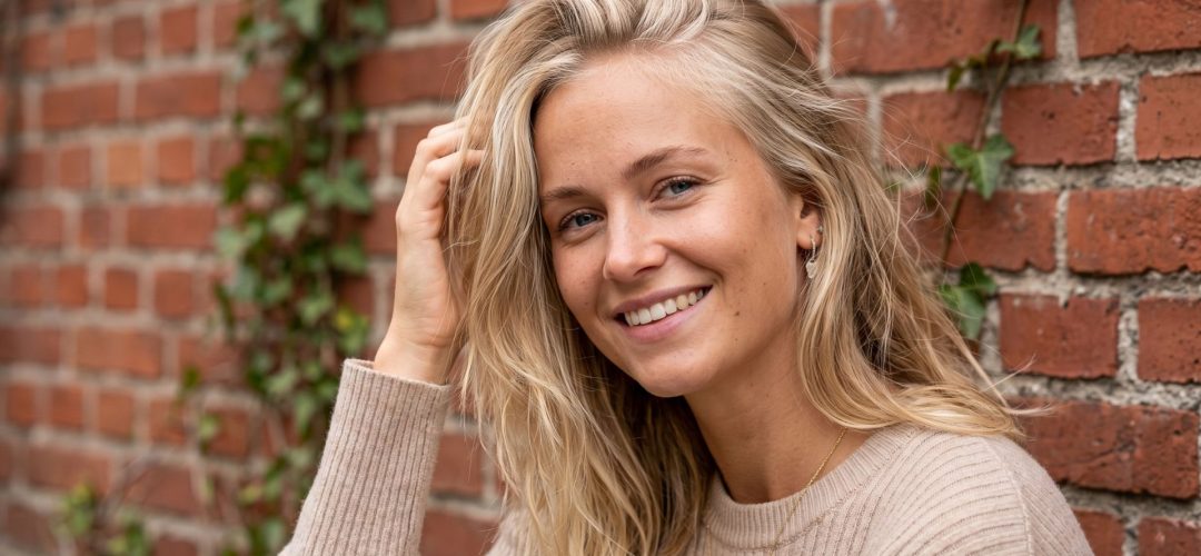 Primo piano di una modella con tecnica Scandinavian Hairline su capelli biondi, posa naturale davanti a un muro di mattoni rossi.