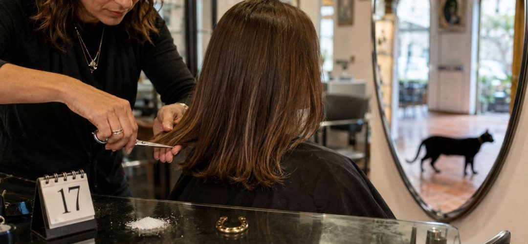 Parrucchiera taglia i capelli a una cliente in salone con calendario 17, sale, ferro di cavallo e gatto nero sullo sfondo.